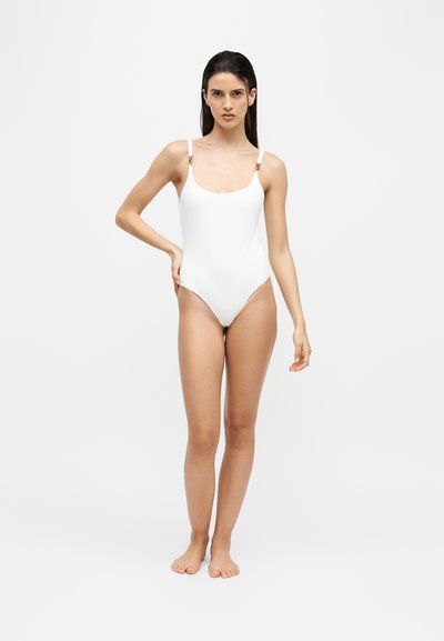 Femme debout pieds nus portant un maillot de bain une pièce blanc avec de fines bretelles et des éléments dorés sur un fond clair uni.