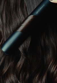 Piastra per capelli nera con un design elegante e una finitura opaca posata su capelli ondulati castano scuro; linee pulite e superficie liscia visibili.