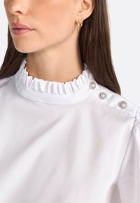 Chemisier blanc avec un col haut à volants, comportant trois boutons décoratifs à l'épaule. Texture lisse, design simple et sans motifs.