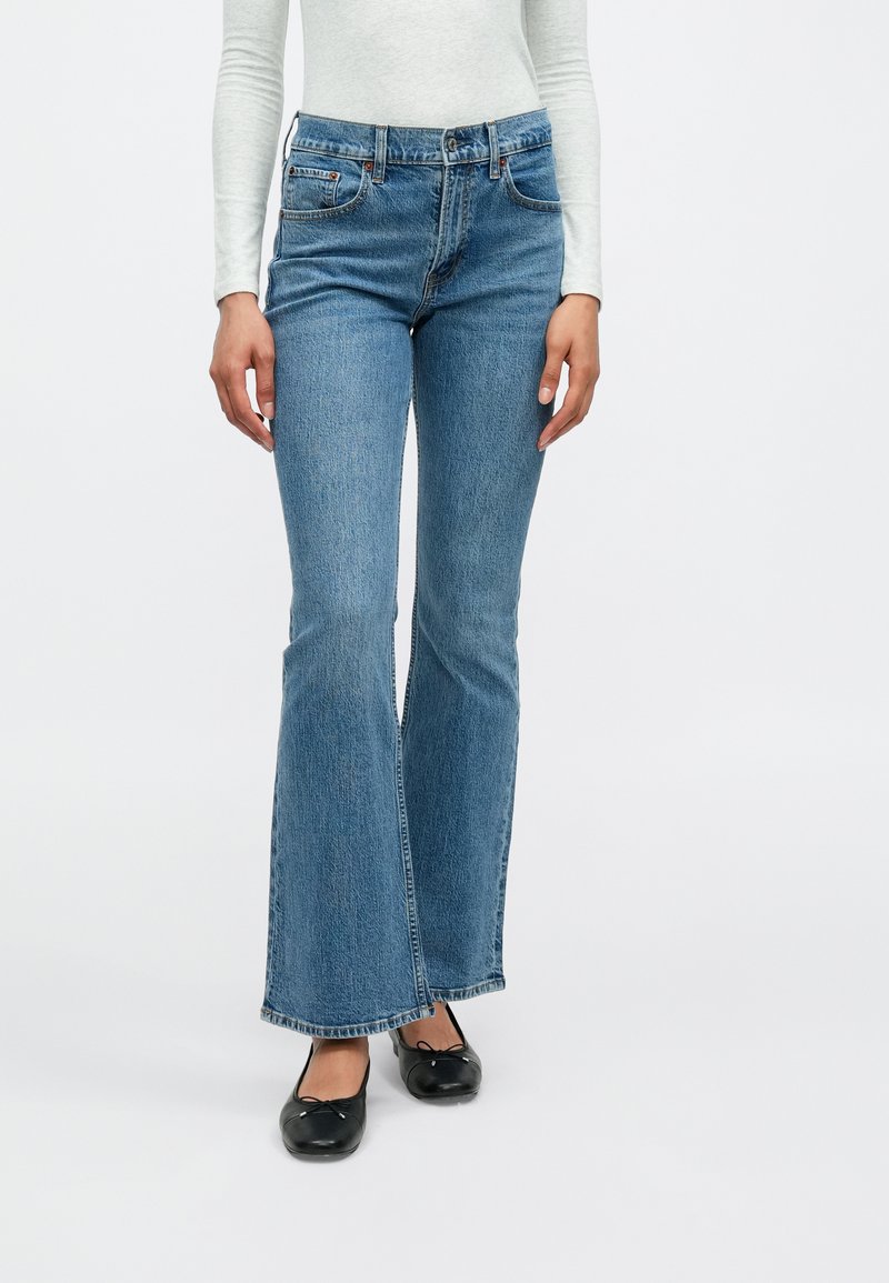 Kavbojke iz modrega denima z razširjenimi hlačnicami in srednje visokim pasom, z velikimi sprednjimi žepi in rahlo opranim videzom, kombinirane s črnimi balerinkami.