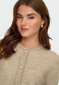 Beżowy kardigan w prążki z perłowymi guzikami, okrągłym dekoltem i miękką teksturą. Modelka ma na sobie złote kolczyki w kształcie kół i falujące, średnio brązowe włosy.