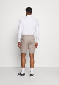 Pantaloni corti color beige chiaro con tessuto testurizzato, vita elasticizzata e due tasche laterali, abbinati a una camicia bianca a maniche lunghe e sneakers nere.
