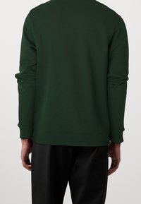 Sudadera verde oscuro con mangas largas y puños acanalados, con cuello redondo y textura suave, combinada con pantalones negros.