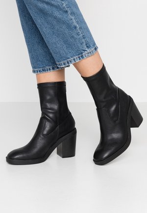 Person iført sorte ankelstøvler med hæl, kombineret med cropped blå denimjeans foran en ensartet baggrund.