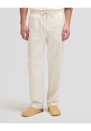 Mężczyzna ubrany w kremowe spodnie z sznurkiem i przednimi kieszeniami naszywanymi oraz w brązowe wsuwane buty, stojący na jednolitym tle.