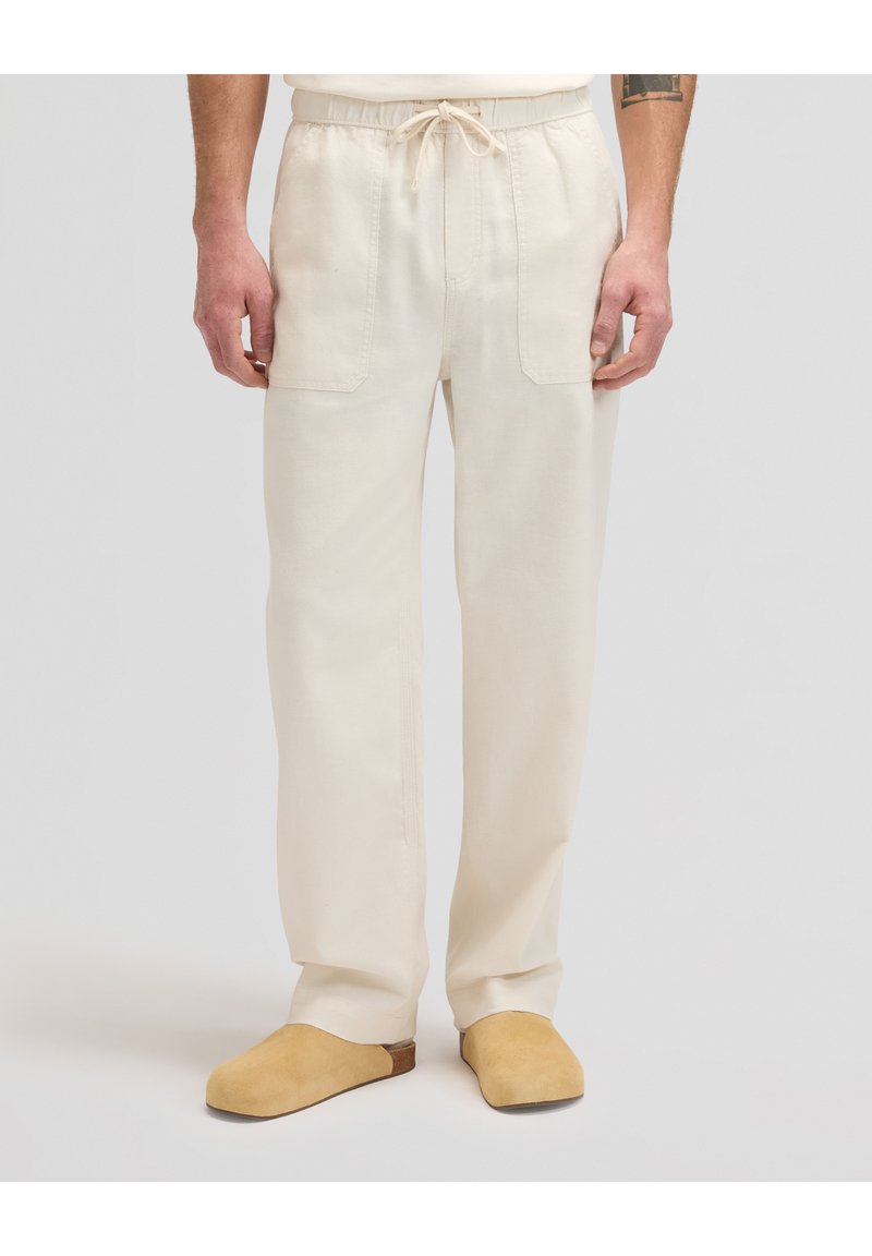 Άνδρας που φοράει μπεζ παντελόνι με κορδόνι στην μέση, εμπρόσθιες τσέπες και ταμπά παπούτσια χωρίς κορδόνια, στέκεται μπροστά σε απλό φόντο.