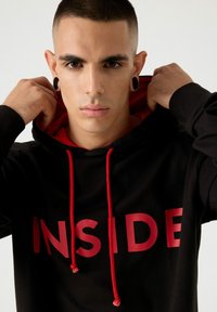 Sudadera negra con detalles rojos y un gran texto "INSIDE". Presenta un forro interior rojo y cordones. Fabricada con una tela suave y con un corte relajado.