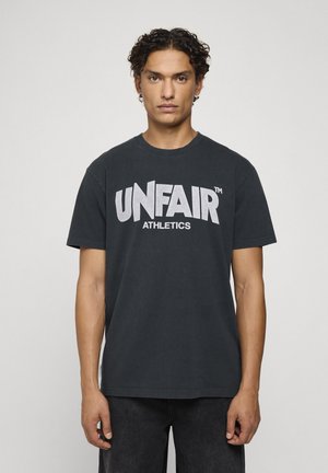 Jeune homme aux cheveux bouclés portant un t-shirt noir avec un grand texte blanc "UNFAIR ATHLETICS" sur un fond uni.