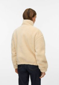 Beige jacka i fuskpäls med hög krage, sjunkna axlar och ribbade muddar, med en mjuk, strukturerad yta och avslappnad passform.