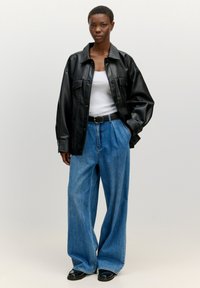 Schwarze Kunstleder-Hemdjacke, weißes Tanktop, weite blaue Jeans, getragen mit einem schwarzen Gürtel. Details umfassen große Taschen und eine glatte Textur.