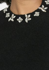 Čierny pletený sveter s okrúhlym výstrihom zdobeným jasnými klenotovými kvetinovými akcentmi. Hladká textúra s priliehavým strihom.