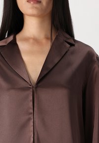 Brown satin shirt featuring a deep V-neck, pointed collar, and a smooth texture. The fabric has a slight sheen and falls gently on the body.