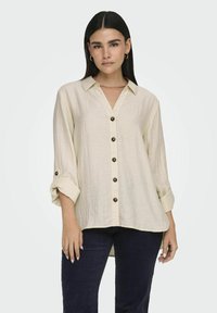 Beige, blouse met knopen van textuurstof; beschikt over een kraag, opgerolde mouwen en donkere knopen aan de voorkant.