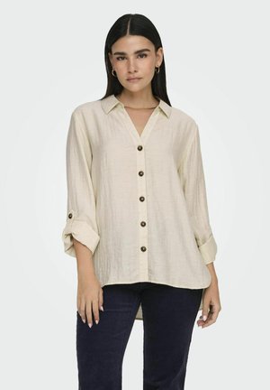 Camicia beige con bottoni realizzata in tessuto testurizzato; presenta un colletto, maniche arrotolate e bottoni scuri lungo la parte anteriore.