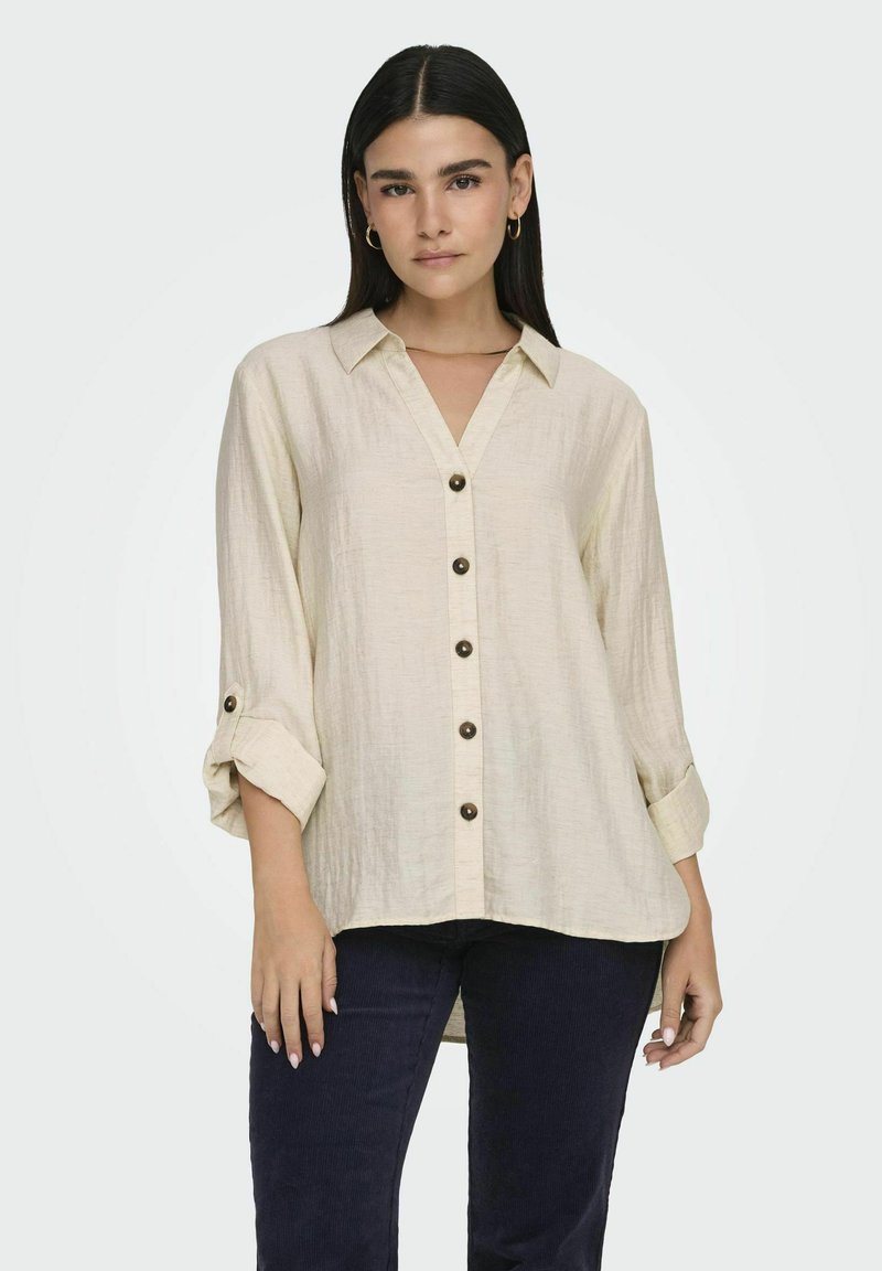 Beige, blouse met knopen van textuurstof; beschikt over een kraag, opgerolde mouwen en donkere knopen aan de voorkant.