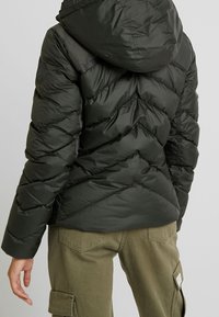 Doudoune en nylon vert foncé avec couture en chevron, capuche et écusson logo sur l'épaule gauche. Associée à un pantalon cargo olive.