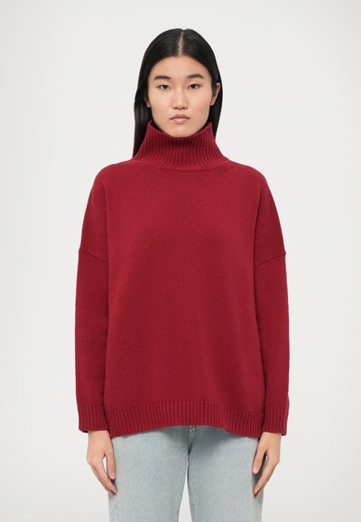 Pull à col roulé rouge, côtelé, avec une coupe décontractée et des épaules tombantes. Texture douce, ourlet avec une finition côtelée subtile, porté sur un jean clair.