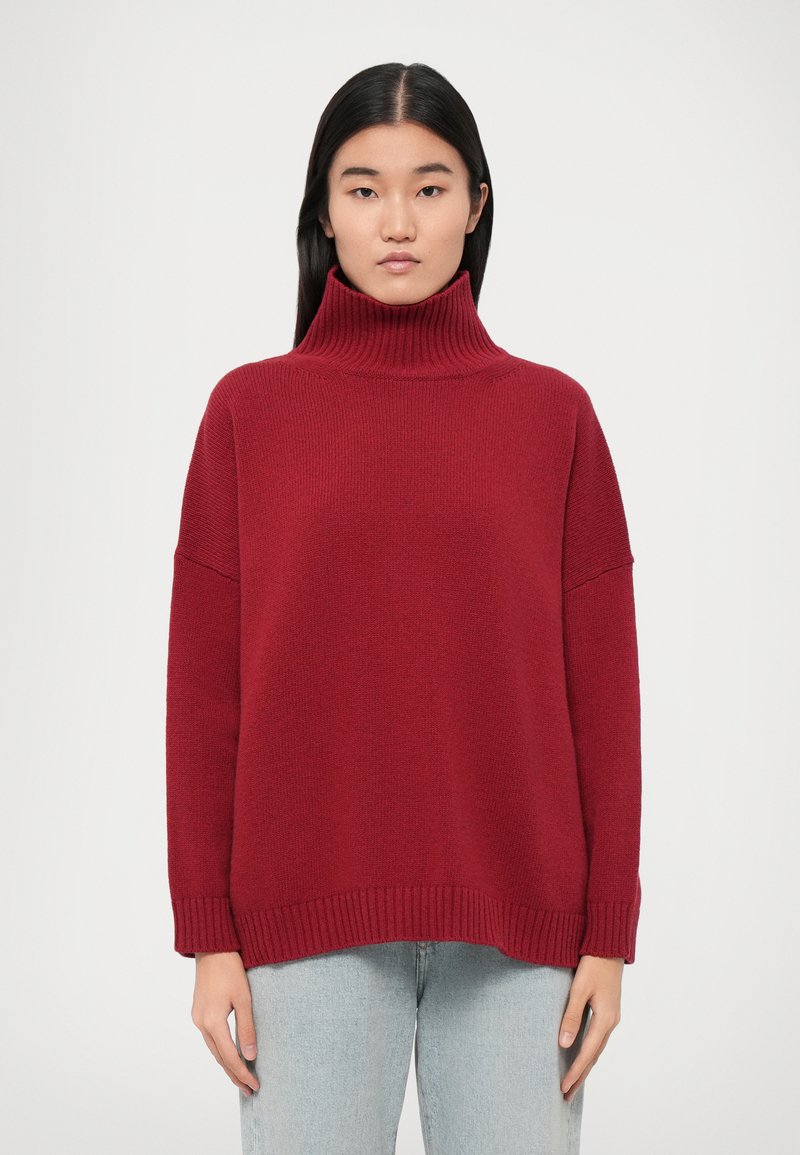 Pull à col roulé rouge, côtelé, avec une coupe décontractée et des épaules tombantes. Texture douce, ourlet avec une finition côtelée subtile, porté sur un jean clair.