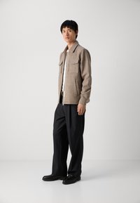 Chaqueta de cremallera a cuadros en marrón y beige, con dos grandes bolsillos en el pecho, combinada con pantalones negros holgados y zapatos negros planos.