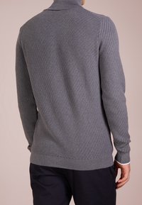 Šedý pletený pulovr s vysokým límcem, s vzorem rybí kosti, ribbed manžetami a hladkou texturou, nošený s tmavými kalhotami.