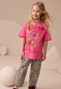 Ružové tričko s krátkym rukávom s motívom medveďa držiaceho jahodu. Kombinované s béžovými nohavicami s leopardím vzorom a bordovými topánkami. Jednoduché pozadie.