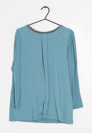Haut à manches longues bleu clair avec un dos plissé. Il présente un col rond agrémenté d'accents métalliques. Fabriqué dans un tissu doux et lisse.