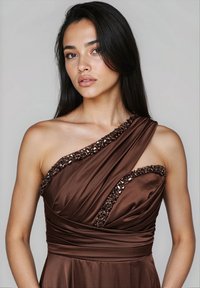 Giovane donna con lunghi capelli scuri, che indossa un abito marrone a spalla unica con bordature di perline, trucco neutro, che guarda leggermente di lato su uno sfondo semplice.