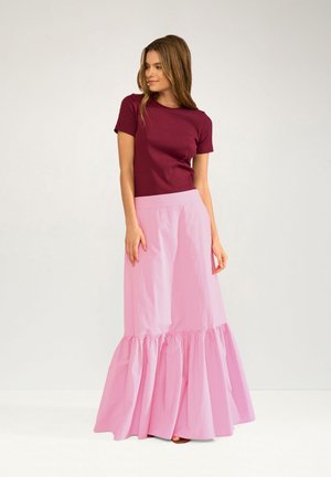 Vrouw die een bordeauxrode top met korte mouwen en een lange lichtroze rok met ruches aan de zoom draagt, staand tegen een effen lichte achtergrond.