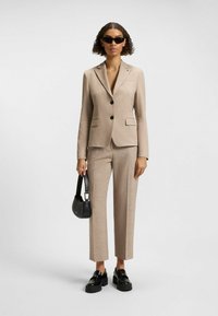 Lichtbeige op maat gemaakt pak met een getailleerde blazer en rechte broek. Zwarte knopen, een enkele borstzak en textielstof. Zwarte tas en zonnebril.
