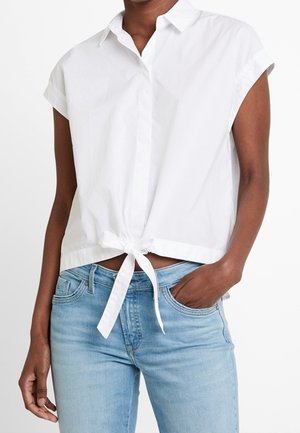 Frau trägt weiße Kurzarmbluse mit vorderer Schleife und hellblaue hoch taillierte Jeans, Hand in der Tasche.