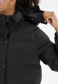 Schwarze Daunenjacke mit hohem Kragen, einem glatten Material, Druckknöpfen und einem dezenten Logo auf der Brust.
