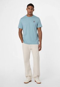 Lyseblå kortærmet bomuld T-shirt med lille logo, kombineret med beige bukser med vidde og hvide sneakers. Enkel, afslappet stil.