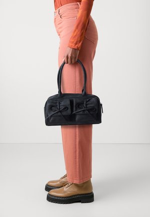 Personne tenant un sac à main noir avec des détails en forme de nœud, portant un pantalon corail, un haut à manches longues orange et des chaussures compensées beige sur un fond uni.