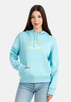 Giovane donna con lunghi capelli scuri che indossa una felpa con cappuccio azzurro chiaro con la scritta gialla "BIDI BADU" e jeans azzurri chiari, in piedi su uno sfondo chiaro.