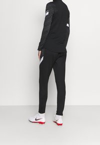 Svart atletisk outfit med en långärmad topp med texturerade ärmar och matchande byxor, accentuerad med vita ränder. Vita fotbollsskor.