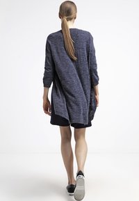 Långärmad, marinblå tröja med avslappnad passform. Har ett lätt, texturerat tyg och en förlängd bakdel, tillsammans med mörka shorts.