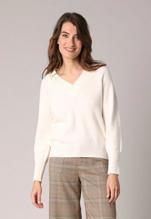 Jeune femme avec des cheveux châtains mi-longs portant un pull blanc à col en V et un pantalon à carreaux marron, souriant à la caméra.