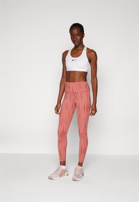 Sutiã desportivo branco com um logótipo preto, combinado com leggings vermelhas estampadas. As leggings apresentam riscas verticais e uma cintura alta. Ténis rosa claro.
