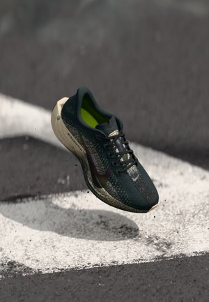 Dunkelgrau Laufschuh mit grünem Innenfutter und weißer Sohle, in der Luft über Asphalt mit weißen Markierungen und Schmutzpartikeln.