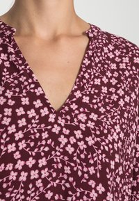Blouse florale bourgogne avec un décolleté en V, ornée de petites fleurs roses et de délicats motifs de branches sur un tissu texturé.
