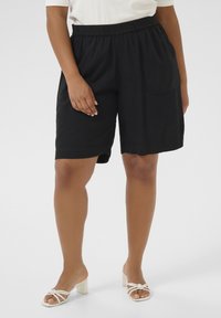 Shorts en lin noir avec une taille élastique et des poches sur les côtés, présentant une coupe décontractée et une texture lisse. Associés à des sandales à talons blanches.
