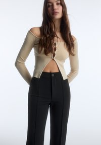 Maglione beige a coste con scollo off-shoulder e chiusura con bottoni frontale, abbinato a pantaloni neri a vita alta con dettaglio di un solo bottone.