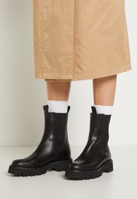 Schwarze Lederstiefeletten mit elastischen Seitenpaneelen, robusten Gummisohlen und Zugschlaufen. Getragen mit weißen Socken und einem beigen Rock.