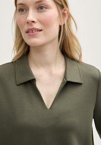 Olive zelená blůza s výstřihem do V a špičatým límcem. Vyrobena z hladkého materiálu; obsahuje ramenní švy a postranní rozparky pro zvýraznění detailu.