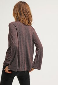 Woman with shoulder-length hair wears a loose, long-sleeve marled knit top and black pants, shown from the back against a plain background.