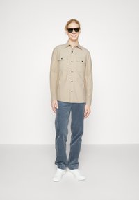 Beige long-sleeve shirt with two chest pockets, button-down front, worn with light blue corduroy pants and white sneakers. Black sunglasses.