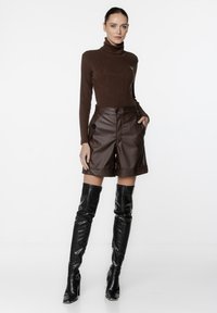 Brązowy sweter z prążkowanym kołnierzem typu turtleneck w połączeniu z brązowymi skórzanymi szortami o wysokim stanie. Modelka nosi błyszczące czarne kozaki za kolano. Gładkie faktury.