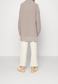 Beige knitted oversized sweater with a high collar, wide sleeves, and ribbed texture, paired with light cream pants and wooden clogs.