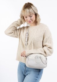 Lys grå håndtaske med et tekstureret panel i hvid pels, buet form og sølv kædedetaljer, båret af en person iført en beige strikket trøje.
