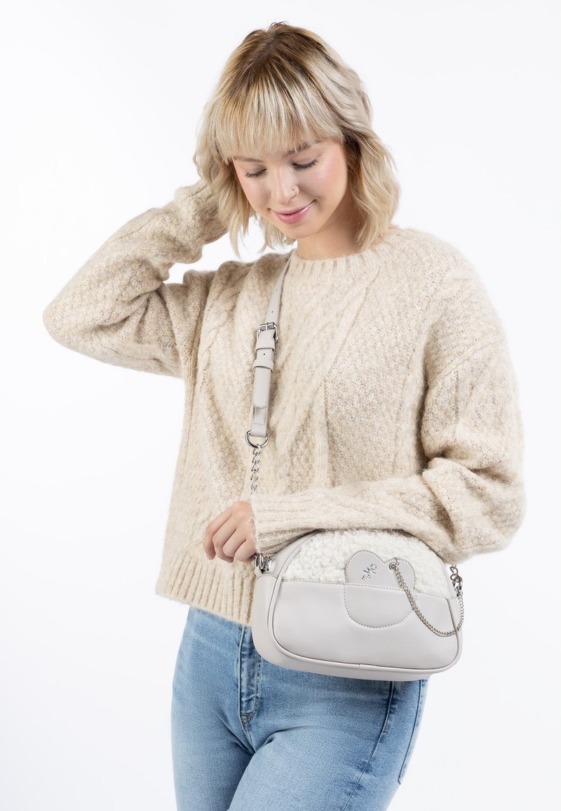 Lys grå håndtaske med et tekstureret panel i hvid pels, buet form og sølv kædedetaljer, båret af en person iført en beige strikket trøje.